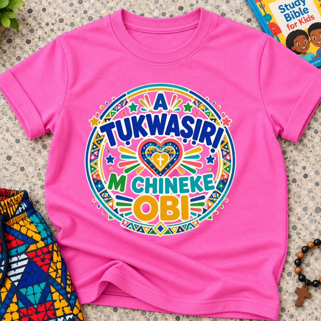 A Tukwasiri M Chineke Obi Igbo-language kids shirt in pink featuring colourful lettering, a heart motif, and circular decorative patterns

